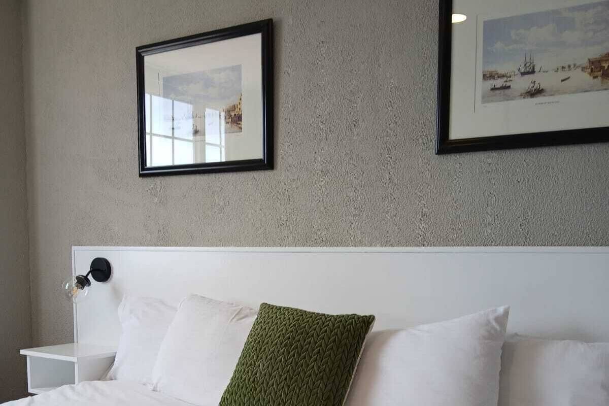 Ein ordentlich bezogenes Bett mit weißen Textilien und einem grünen Zierkissen, ein weißer Nachttisch mit Lampe sowie zwei gerahmte Bilder an der Wand.