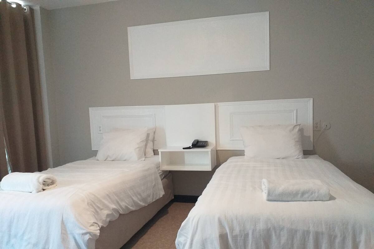 Ein Hotelzimmer mit zwei Einzelbetten, jeweils mit einem Kopfkissen und einem zusammengerollten Handtuch. Dazwischen befindet sich ein Nachttisch mit Telefon und Lampe.