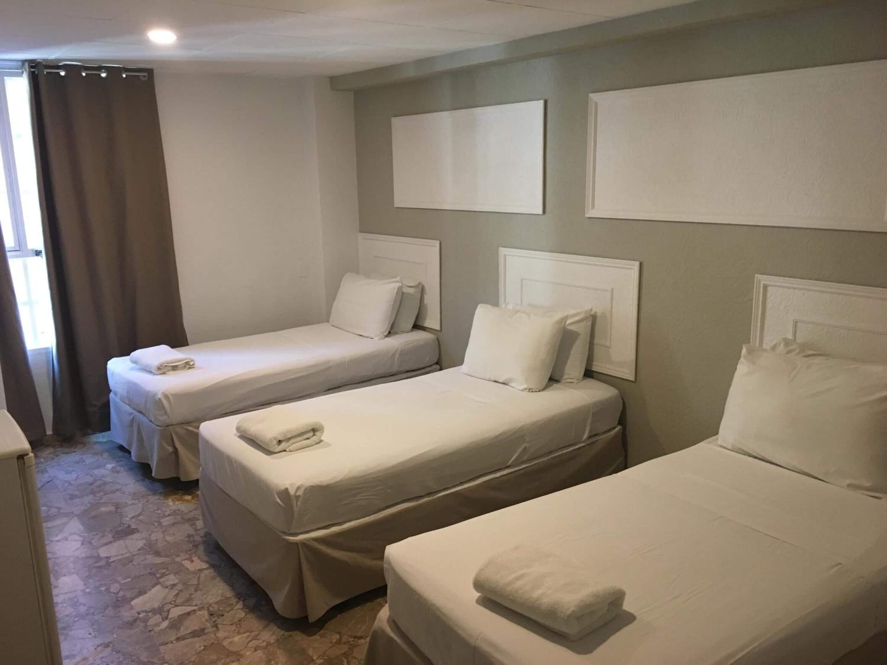 A hotel room with three single beds, each with white linens and pillows, and a small bedside table.