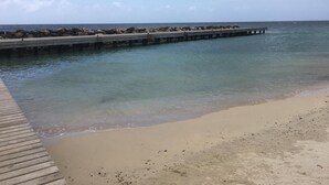 Une plage avec une jetée en bois s’avançant dans des eaux calmes, un rivage sablonneux et un ciel dégagé.