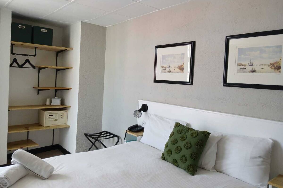 Ein Hotelzimmer mit Bett, Schreibtisch mit Lampe, Stuhl, Ablagefläche mit Aufbewahrungsboxen und zwei gerahmten Bildern an der Wand.