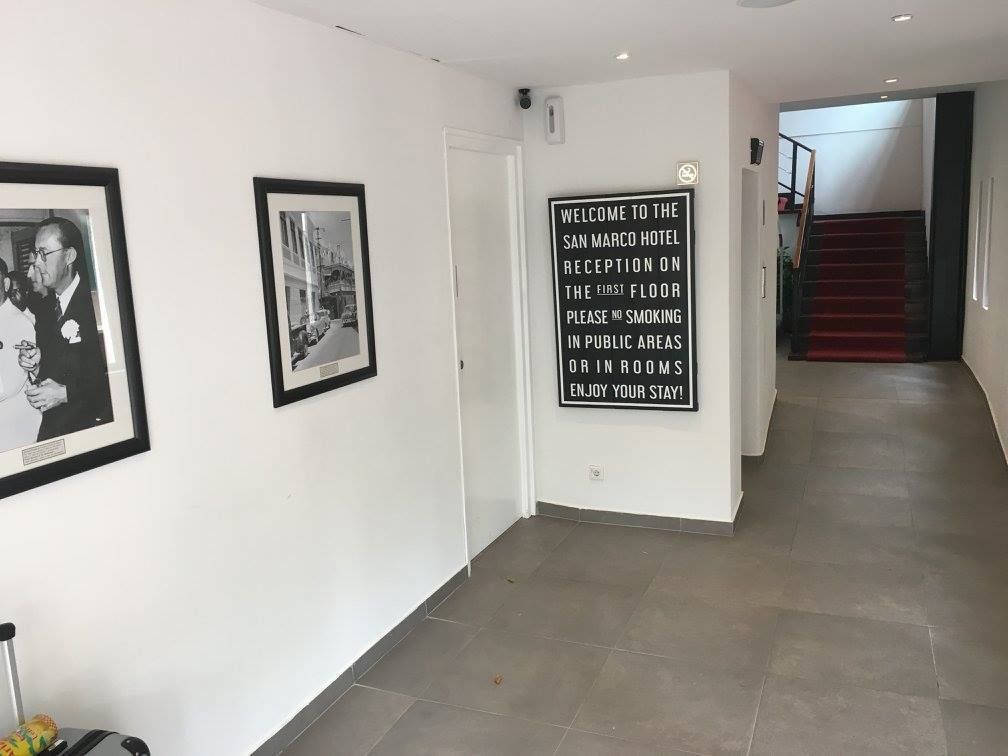 Zona de recepción de un hotel con fotografías enmarcadas en las paredes, un cartel de prohibido fumar y una escalera al fondo.