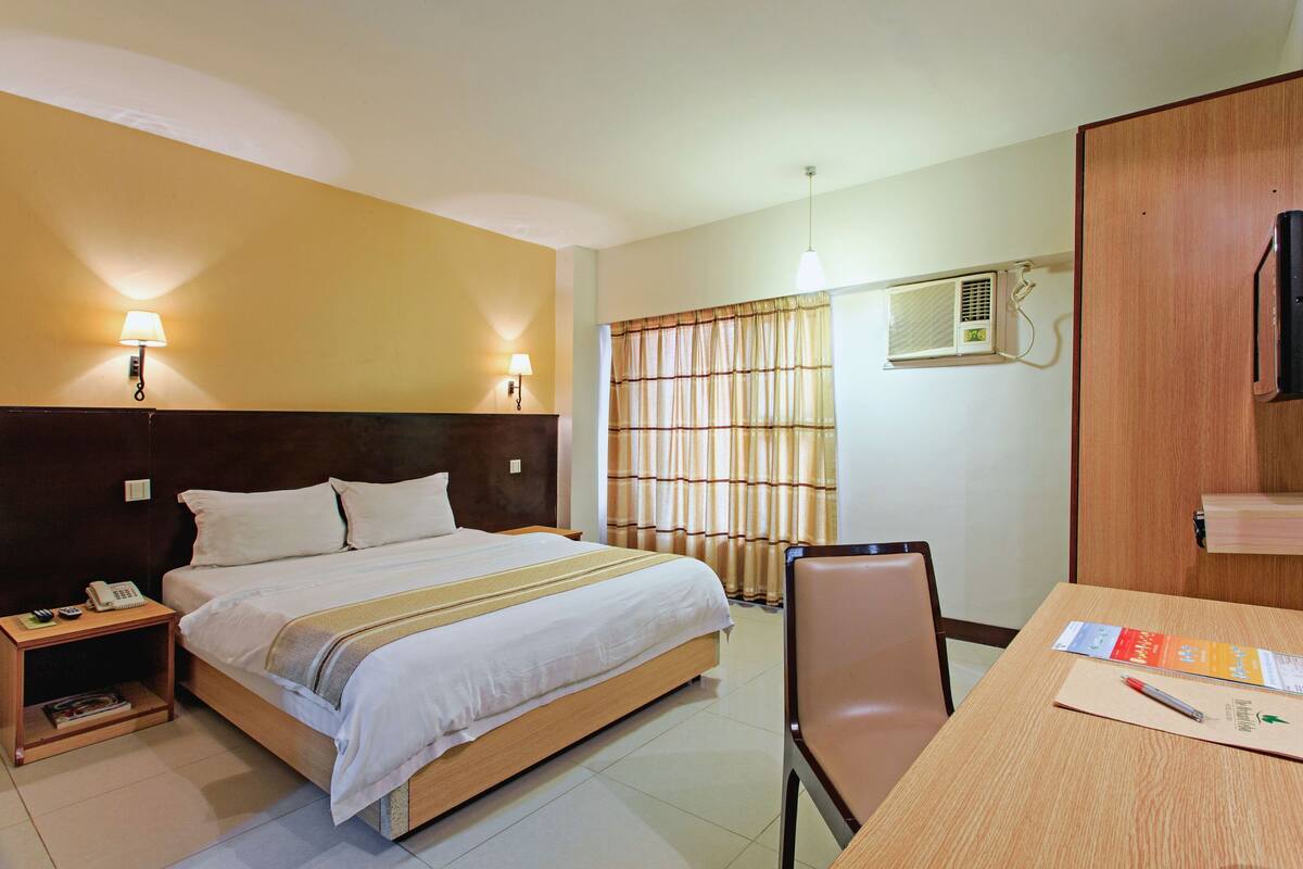 A hotel room with a bed, bedside tables, a desk with a chair, a lamp, an air conditioning unit, and a television.