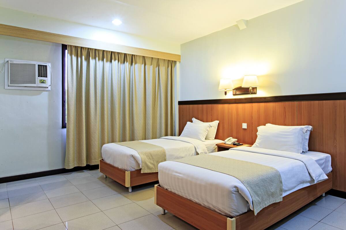 A hotel room with two single beds, a wooden headboard, a bedside table with a lamp, and an air conditioning unit.