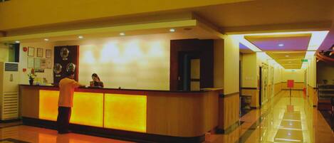A hotel reception area with a reception desk, a receptionist, and a hallway leading to other rooms.