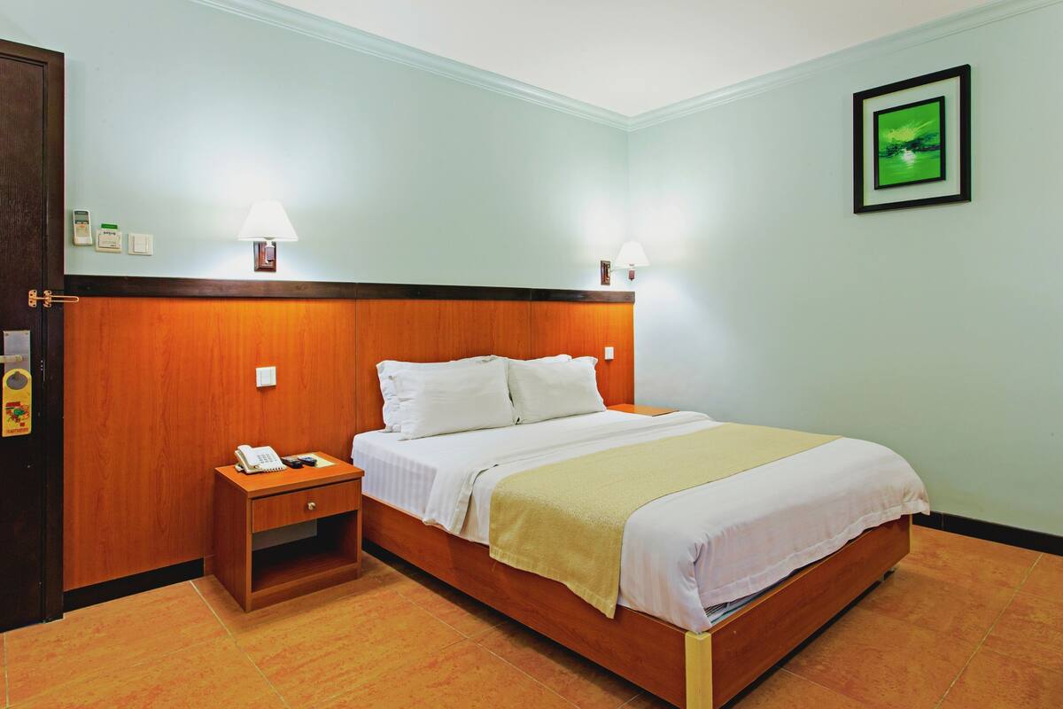 A hotel room with a wooden headboard, a bed with white linens, a bedside table with a phone, and a wall-mounted lamp.