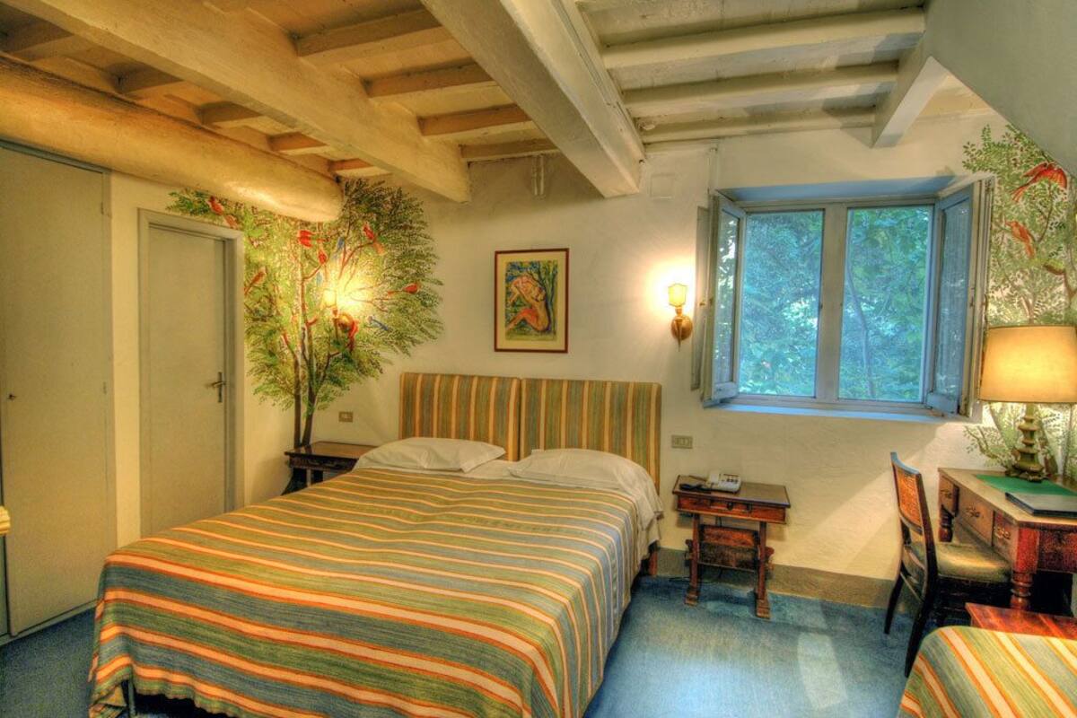 A cozy bedroom with a striped bedspread, a nightstand with a lamp, a chair, and a window with a view of trees.