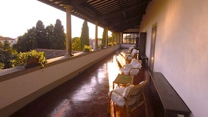 Un espacio de estar techado al aire libre con sillas de mimbre y una mesa larga, con vistas a un jardín y edificios.