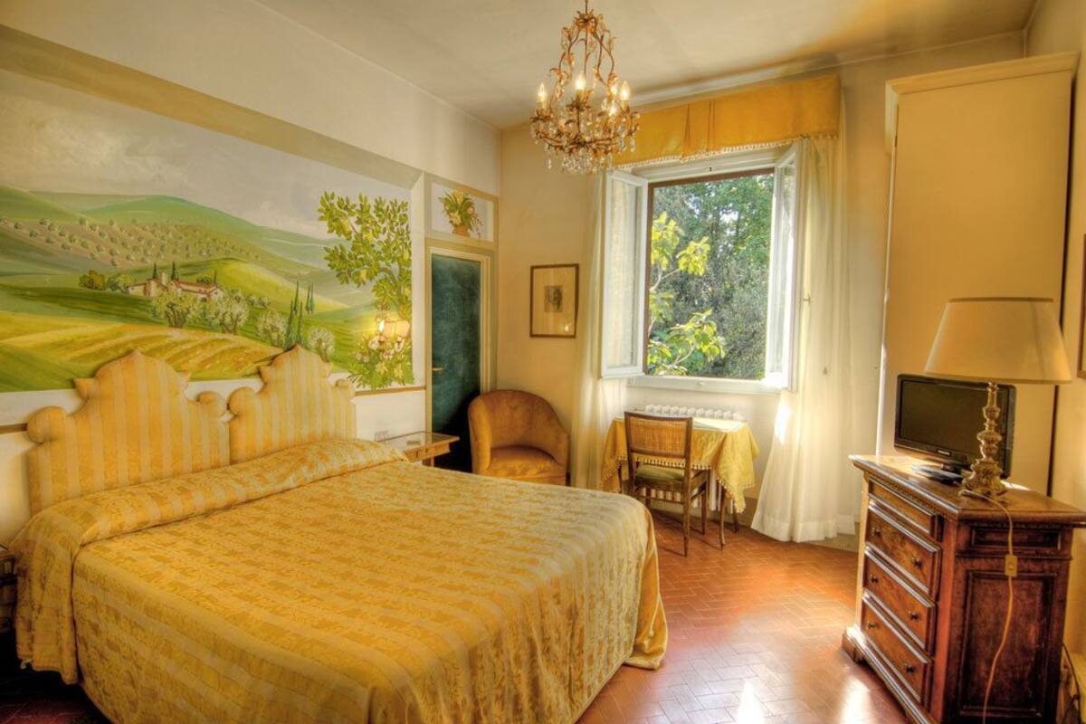 A bedroom with a large bed, a desk with a chair, a television on a dresser, and a window with curtains.