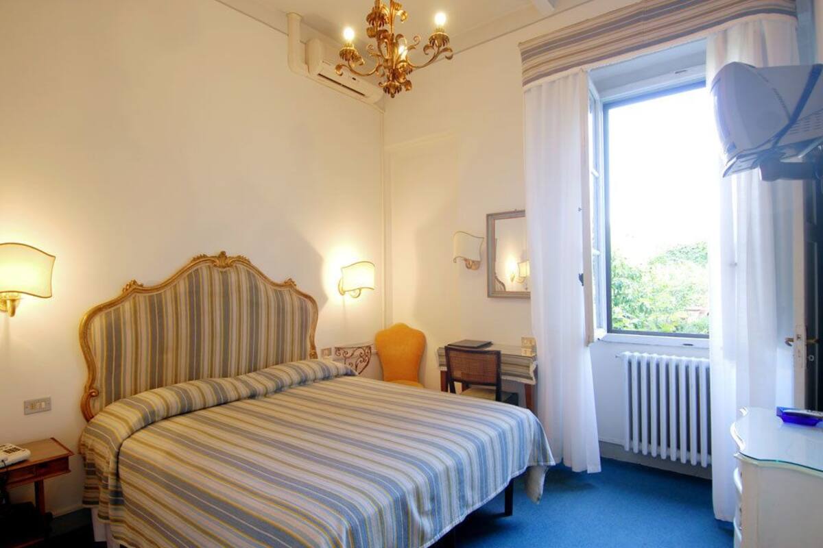 A hotel room with a large bed, a desk, a chair, a window with curtains, and a radiator.