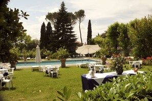 Un espace extérieur avec une piscine, des chaises longues et des parasols, le tout entouré de verdure et d’arbres.