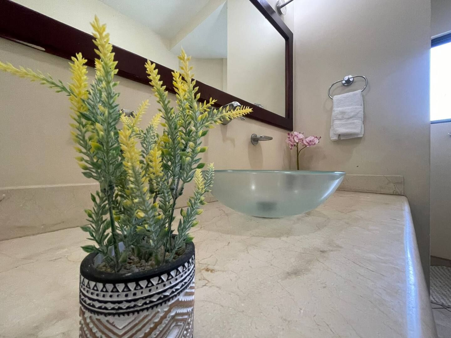 Une salle de bain avec un lavabo en verre, une plante en pot aux fleurs jaunes, un miroir et un porte-serviettes.
