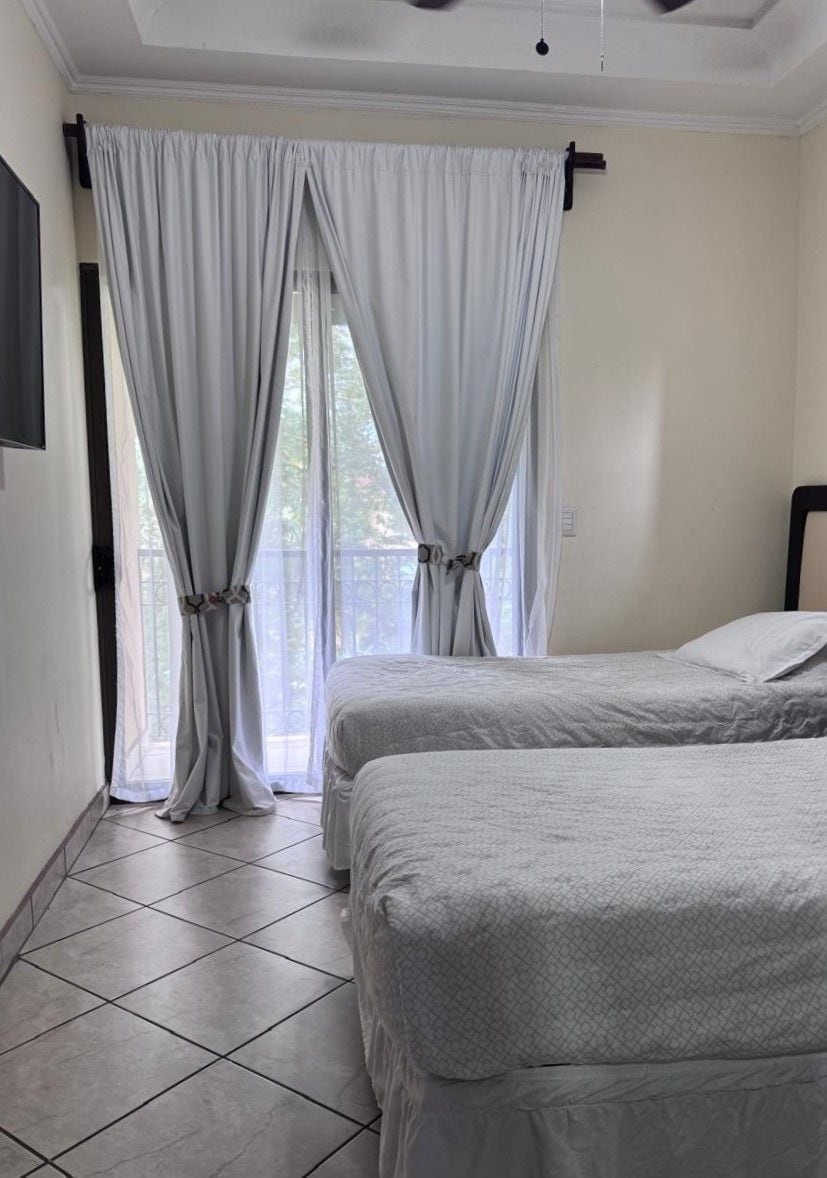 A room with two beds, a window with curtains, and a tiled floor.