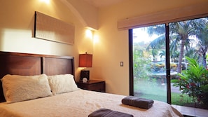 A bedroom with a large bed, a nightstand, a lamp, and a view of a garden through a window.