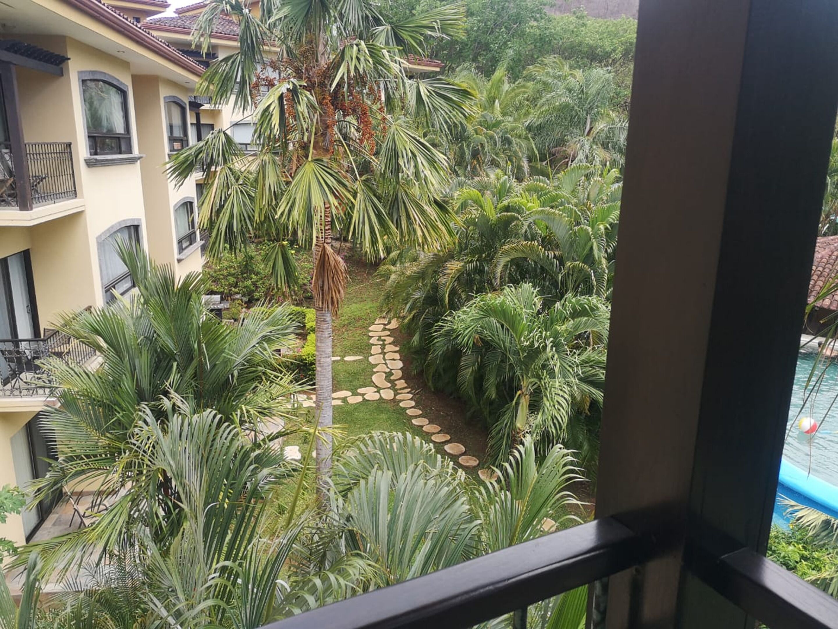 A balcony with a view of a lush garden, a swimming pool, and a building with balconies.