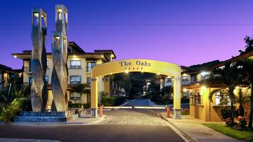 Entrada de un hotel con una escultura moderna, un letrero que dice "The Oaks" y un camino de acceso bien iluminado.