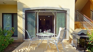 A patio with a round table and chairs, a grill, and a view of the interior through a glass door.