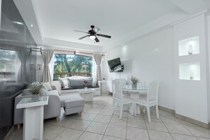 A modern living room with a sofa, ottoman, and dining table, a ceiling fan, and a TV mounted on the wall. - The Oaks Tamarindo Condominiums (Tamarindo)