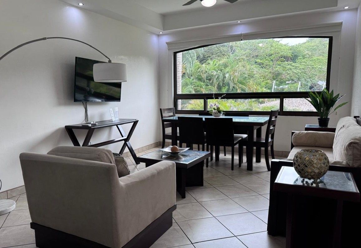 Una sala moderna con un televisor de pantalla plana, un sofá, una mesa de comedor con sillas y un ventanal que ofrece vistas a zonas verdes.