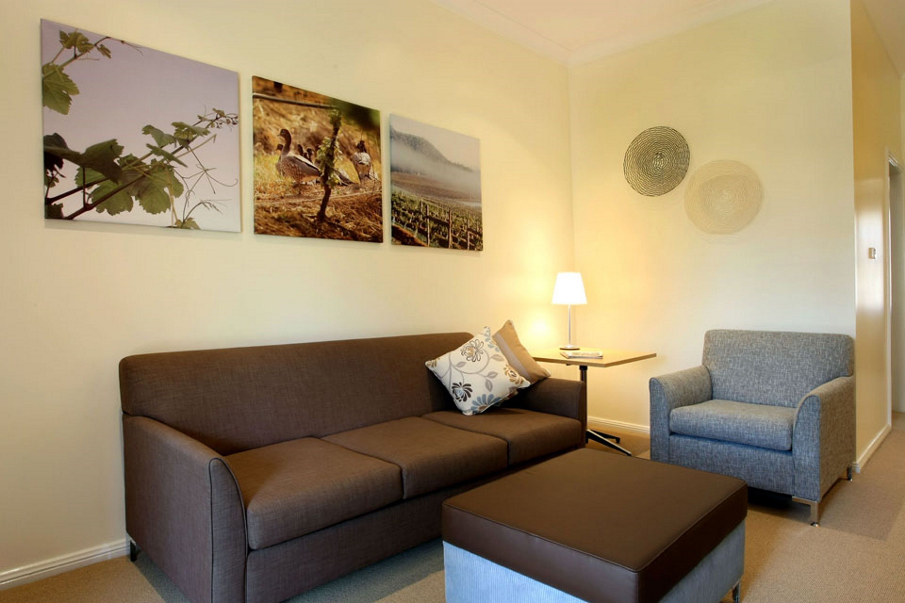Een woonkamer met een bruine bank, een blauwe fauteuil en een klein tafeltje met een lamp. Drie ingelijste foto's hangen aan de muur.
