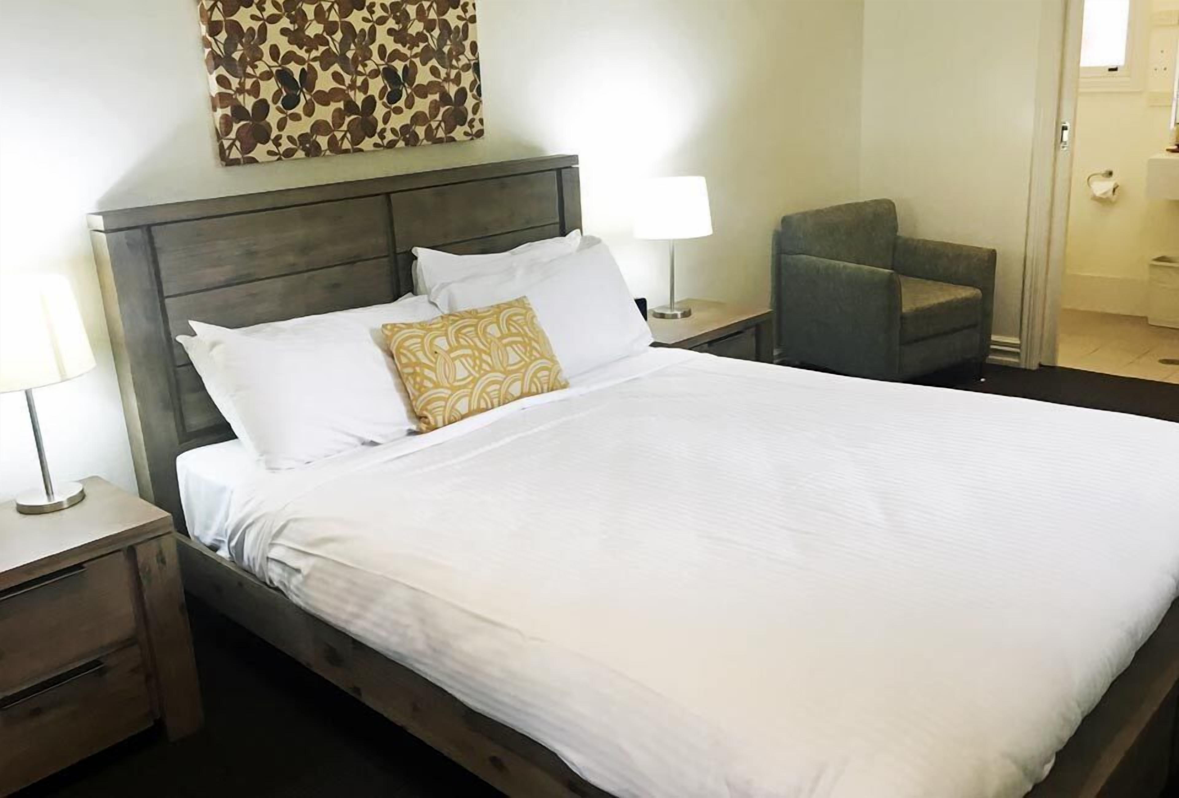 A neatly made bed with white linens and a patterned pillow, a wooden nightstand with a lamp, a grey armchair, and a wall-mounted artwork.