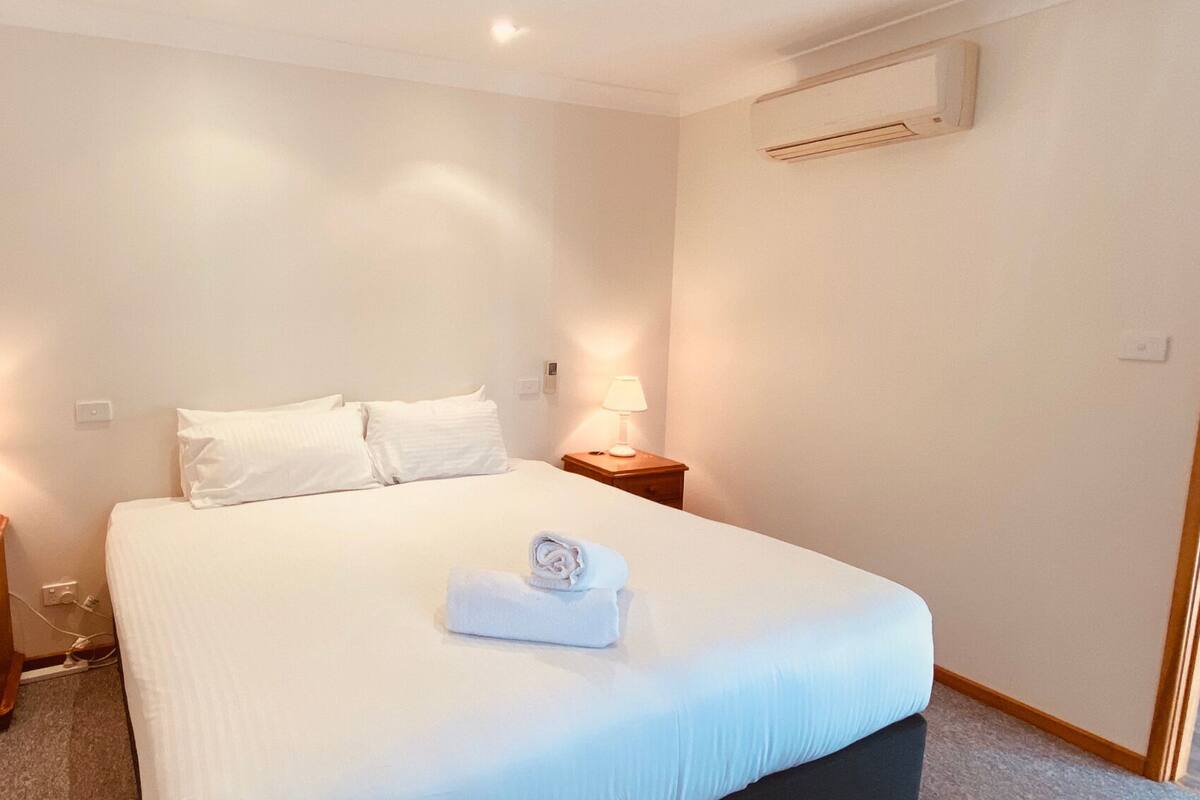 Un lit bien fait, avec du linge de lit blanc et une serviette pliée, deux lampes de chevet, une table de nuit en bois et un climatiseur fixé au mur.
