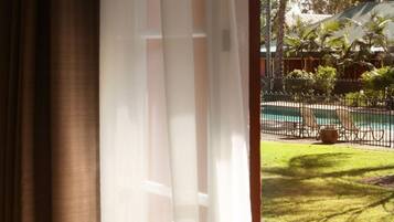 Vista a través de una ventana con cortinas, mostrando una piscina, un área verde y un jardín vallado con palmeras.