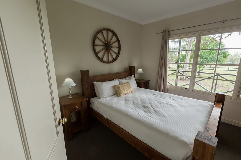 A bedroom with a wooden bed, two bedside tables with lamps, a large window with curtains, and a wooden wheel on the wall.