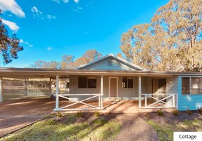 A cottage with a covered porch, a fenced driveway, and a view of trees.