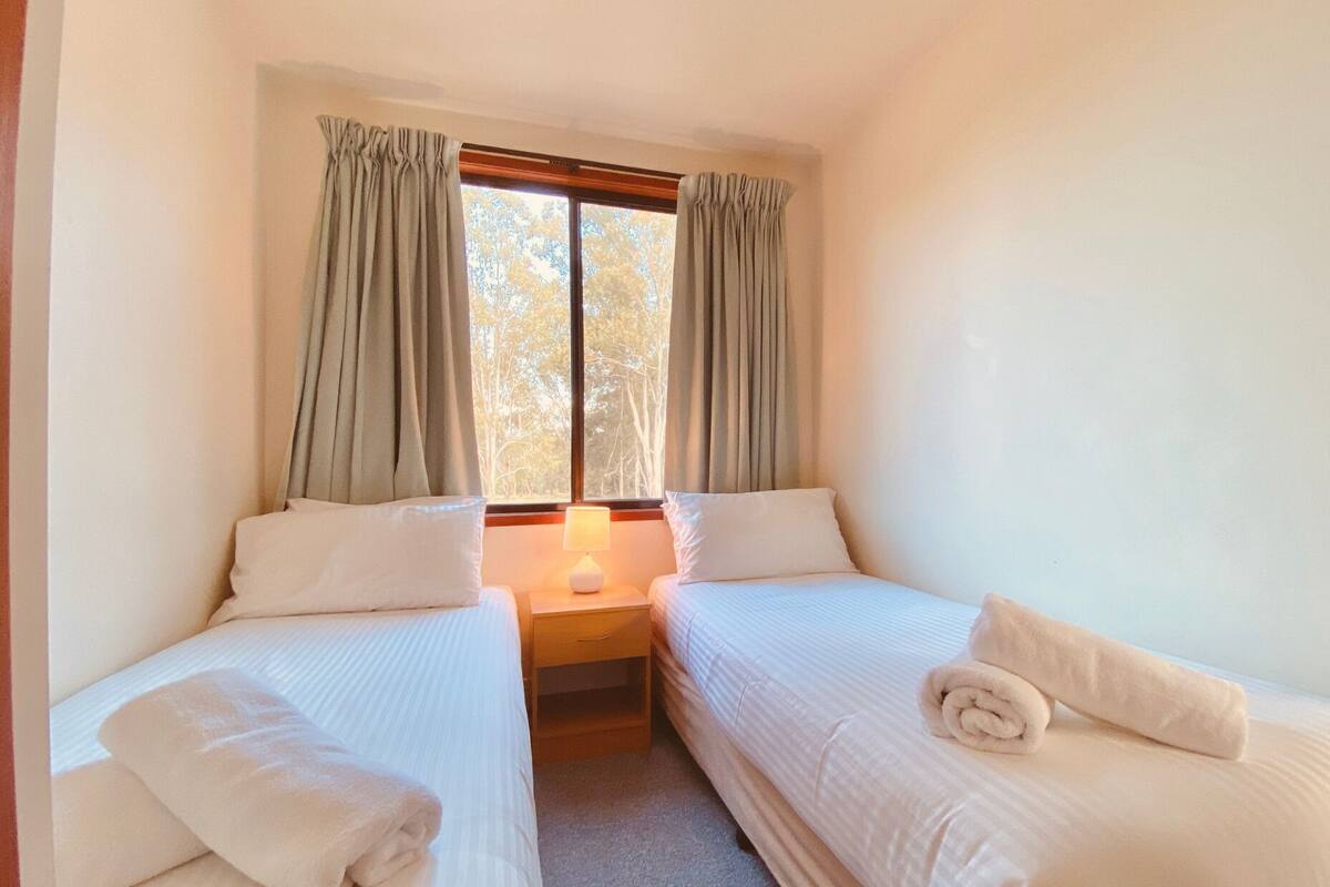 Une petite chambre propre, avec deux lits simples, une table de chevet avec une lampe et une fenêtre avec des rideaux.