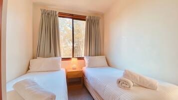 Quarto pequeno e limpo com duas camas de solteiro, uma mesa de cabeceira com abajur e uma janela com cortinas.