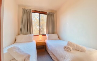 Quarto pequeno e limpo com duas camas de solteiro, uma mesa de cabeceira com abajur e uma janela com cortinas.