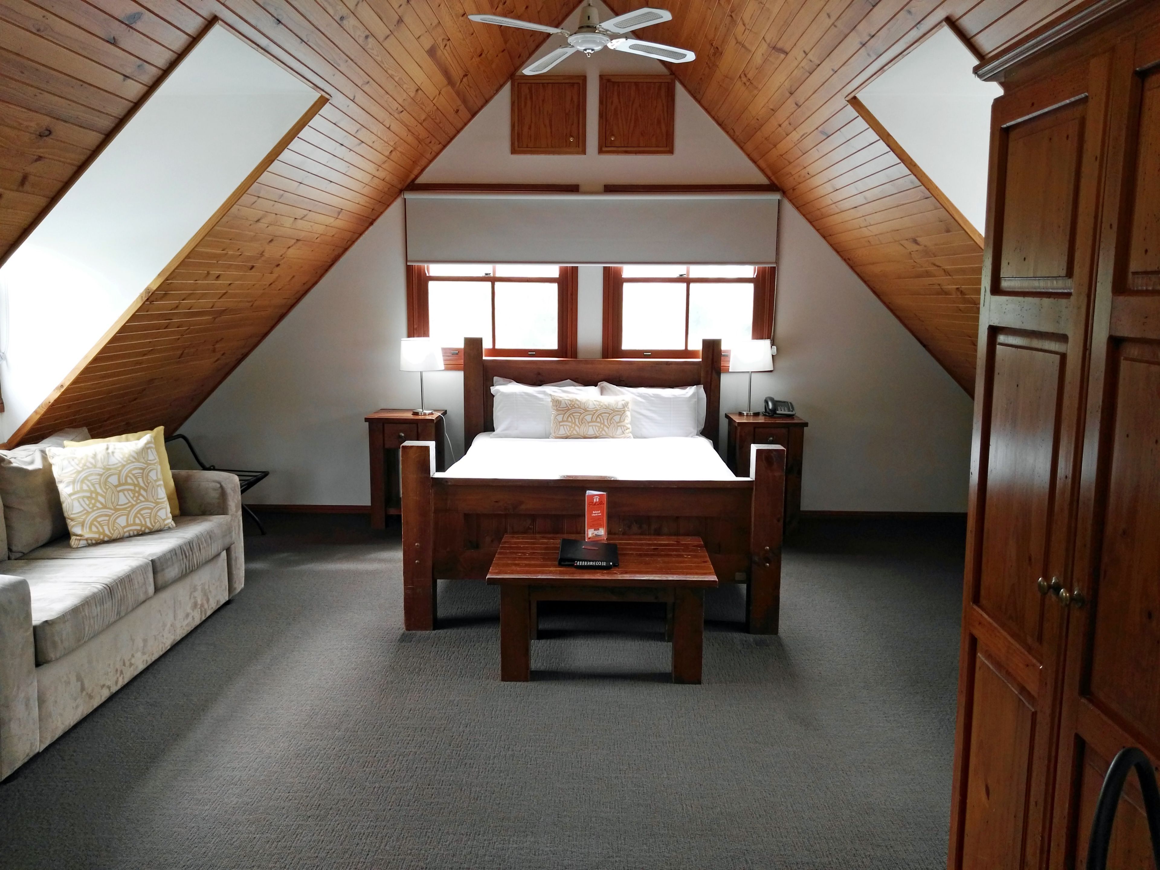A bedroom with a wooden ceiling, a bed with white bedding, a nightstand, a bedside table, a lamp, a wooden wardrobe, and a sofa with patterned cushions.