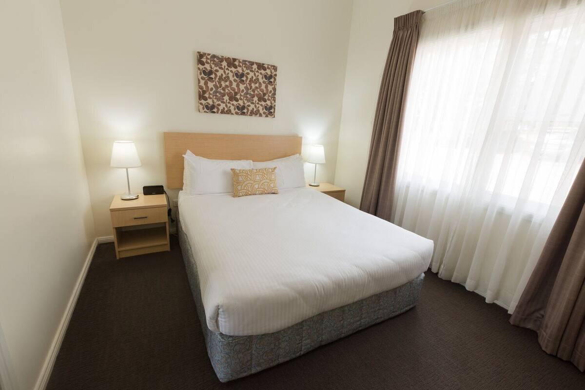 Une chambre d’hôtel avec un grand lit, deux tables de chevet avec des lampes, une tête de lit, une œuvre d’art accrochée au mur et une fenêtre avec des rideaux.