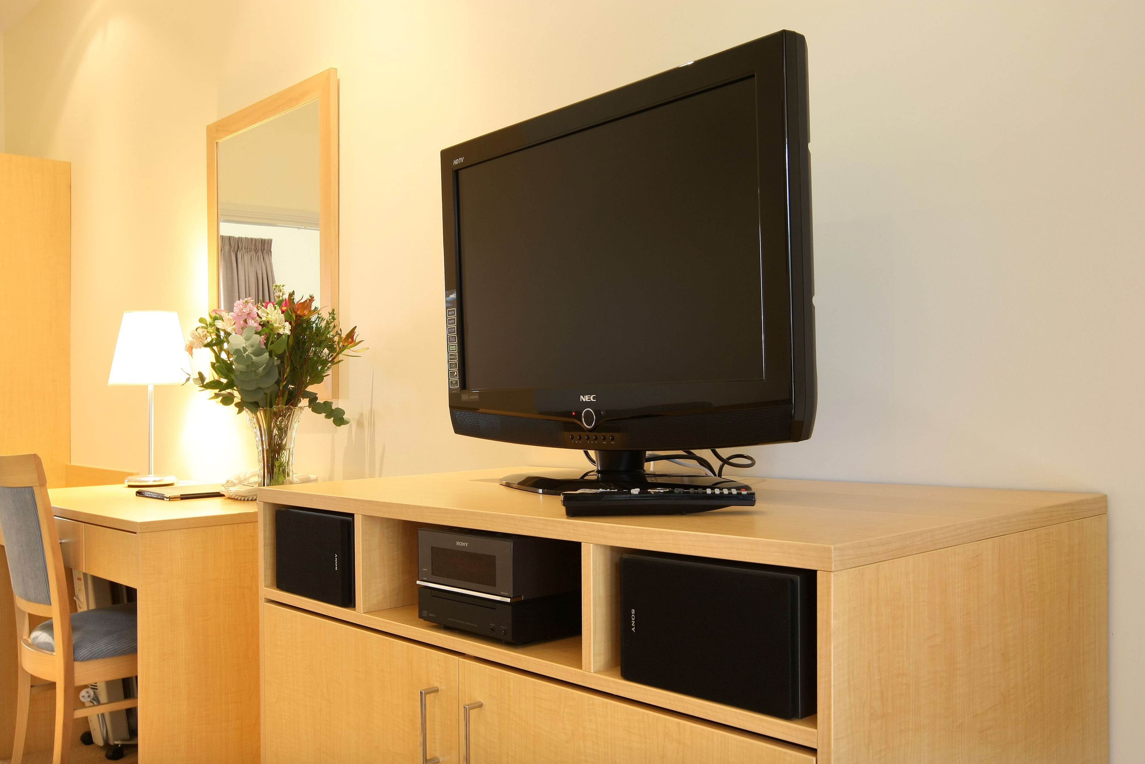 A hotel room with a wooden desk, a flat-screen TV, a Sony audio system, a lamp, and a vase with flowers.