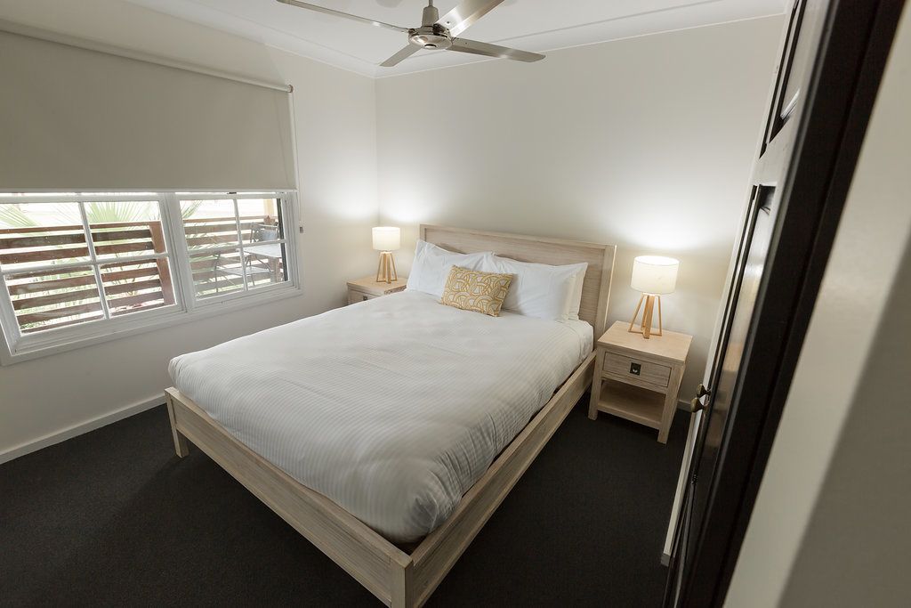 Une chambre à coucher comprenant un lit, deux tables de chevet, un ventilateur de plafond et une fenêtre avec des stores.