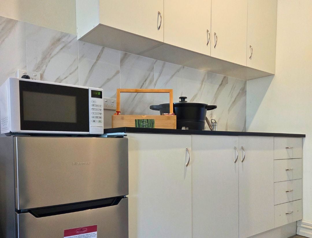 A kitchen with a stainless steel refrigerator, a microwave, and a wooden cutting board with a pot. The kitchen has white cabinets and a marble backsplash.
