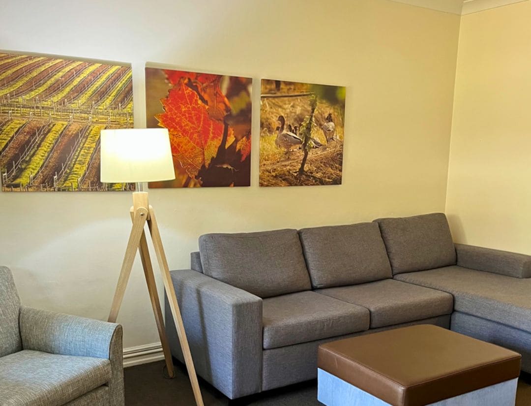 A living room with a grey sofa, a brown ottoman, a small grey armchair, and a floor lamp. There are three framed artworks on the wall.