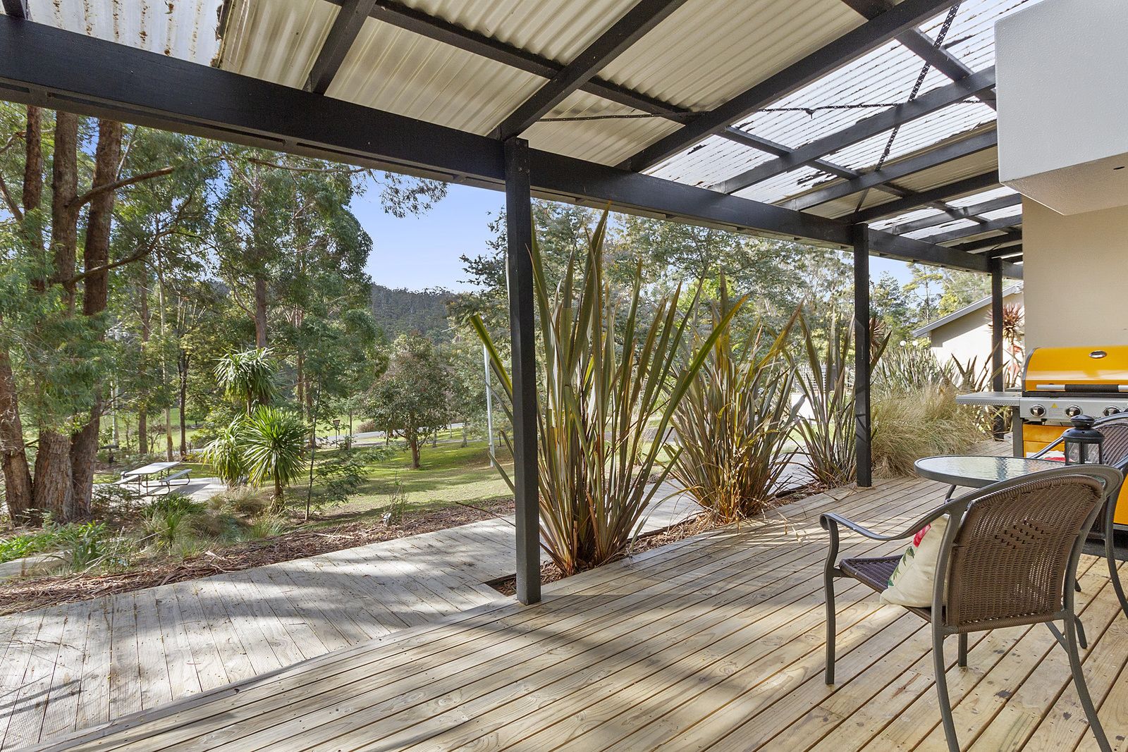A covered outdoor area with a wooden deck, a grill, and a view of a park with trees and a distant mountain.