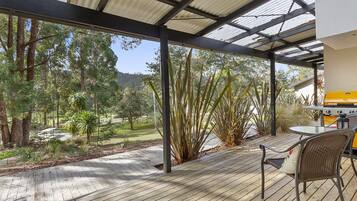 A covered outdoor area with a wooden deck, a grill, and a view of a park with trees and a distant mountain.