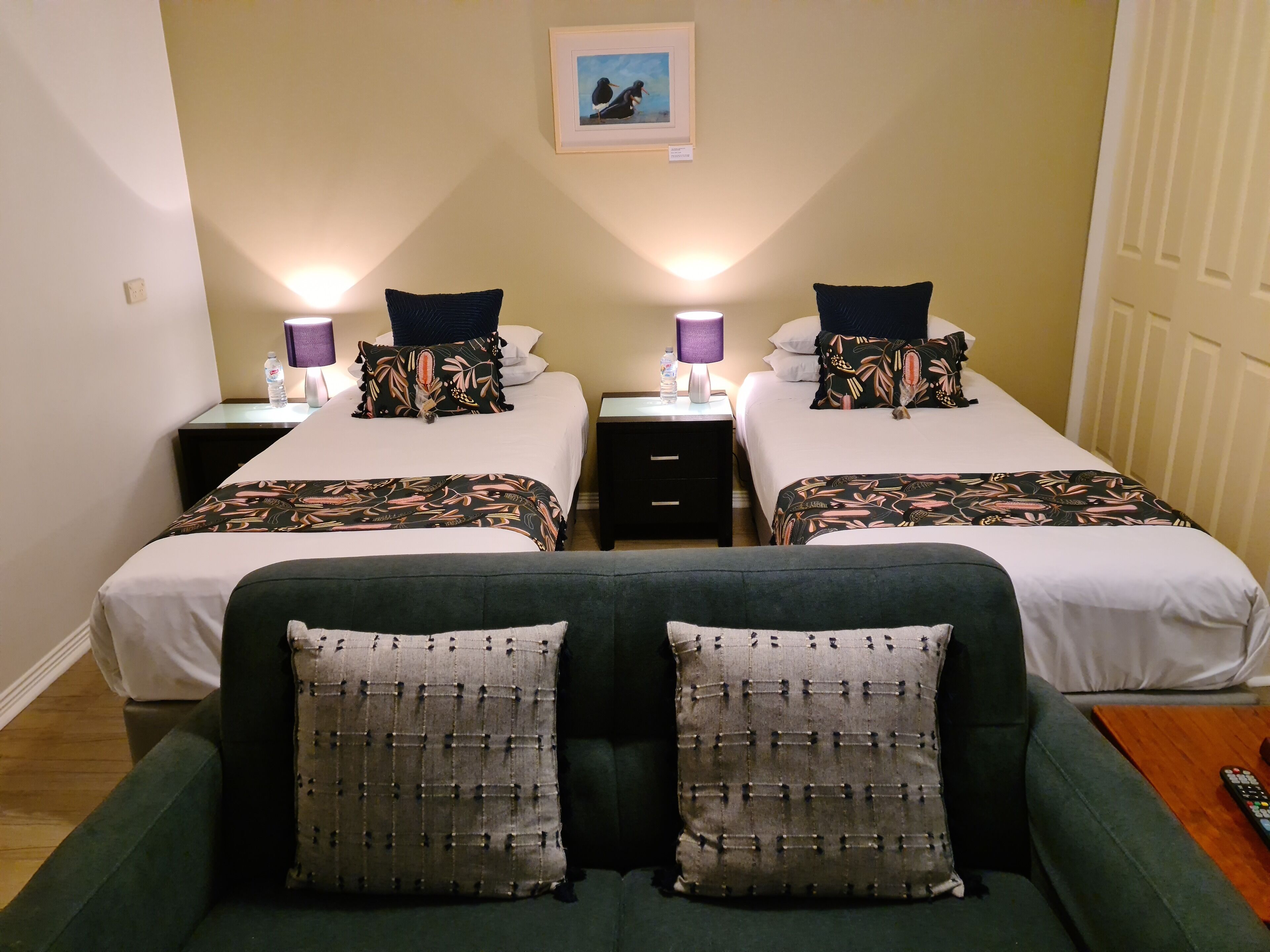 A double room with two beds, each with a patterned bedspread and pillows, two bedside tables with lamps, a framed picture on the wall, and a green sofa with patterned cushions.