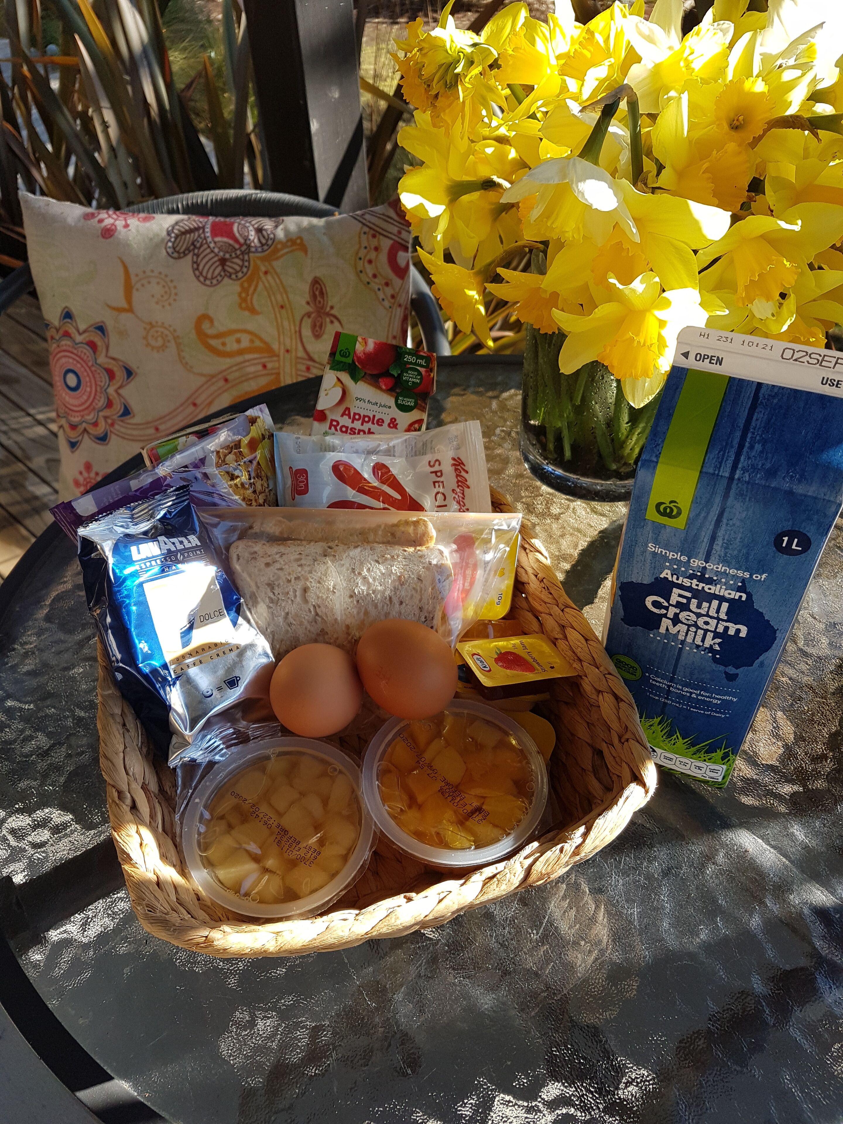 Een mand met etenswaren, een pak melk en een vaas met gele bloemen op een tafel.