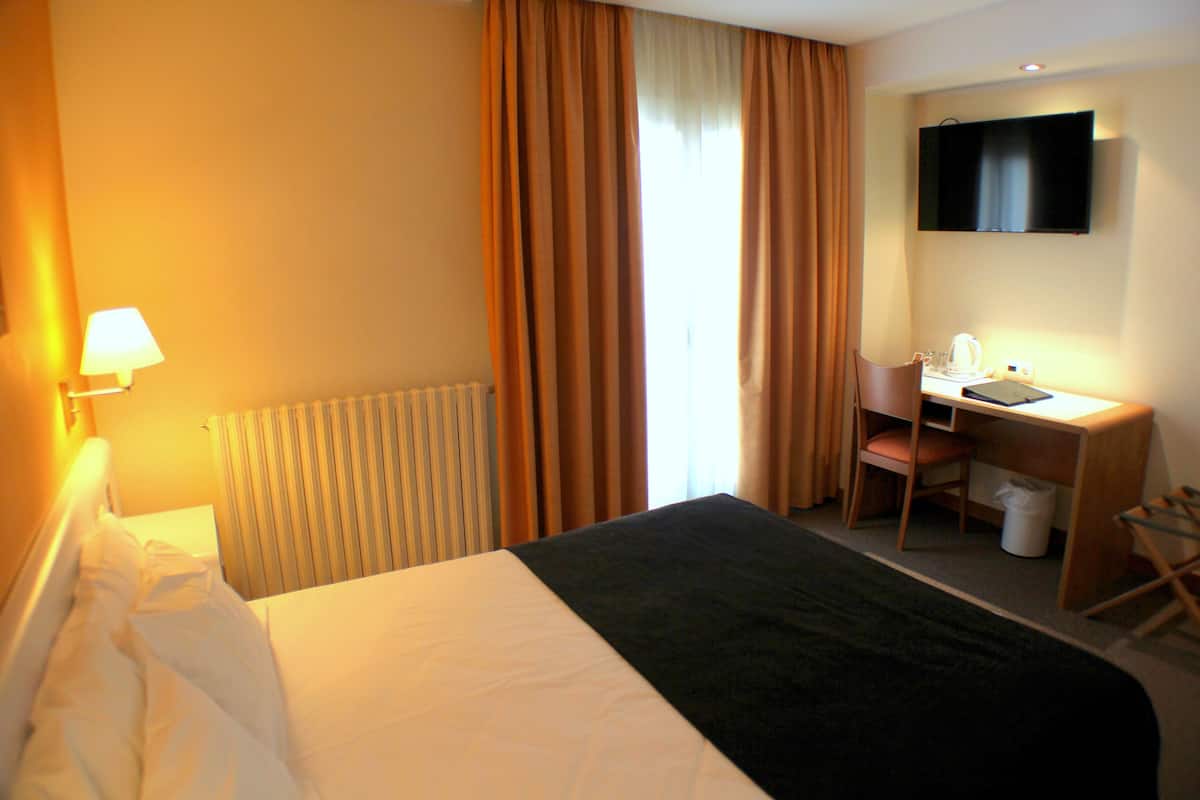 A hotel room with a bed, a desk with a chair, a television, and a window with curtains.