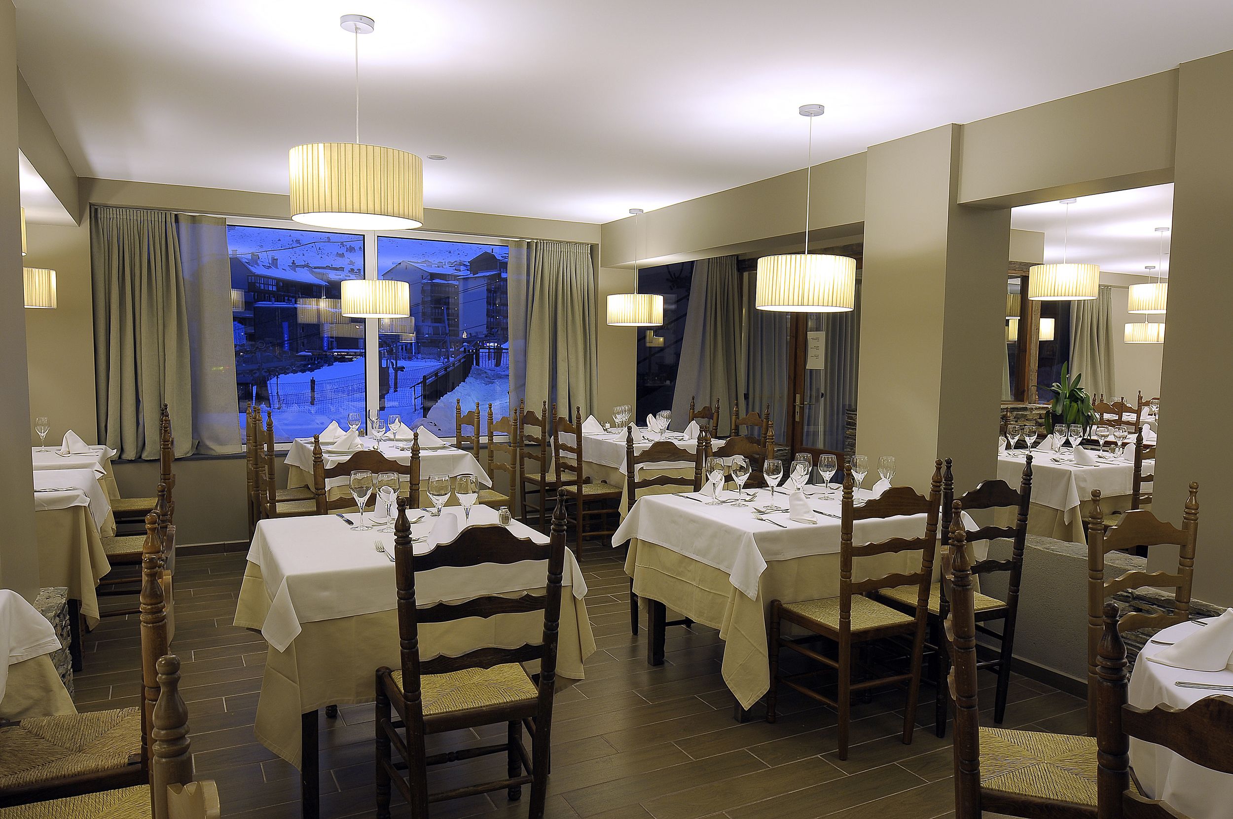 Comedor con mesas dispuestas para una comida, sillas de madera y ventanas grandes con cortinas.