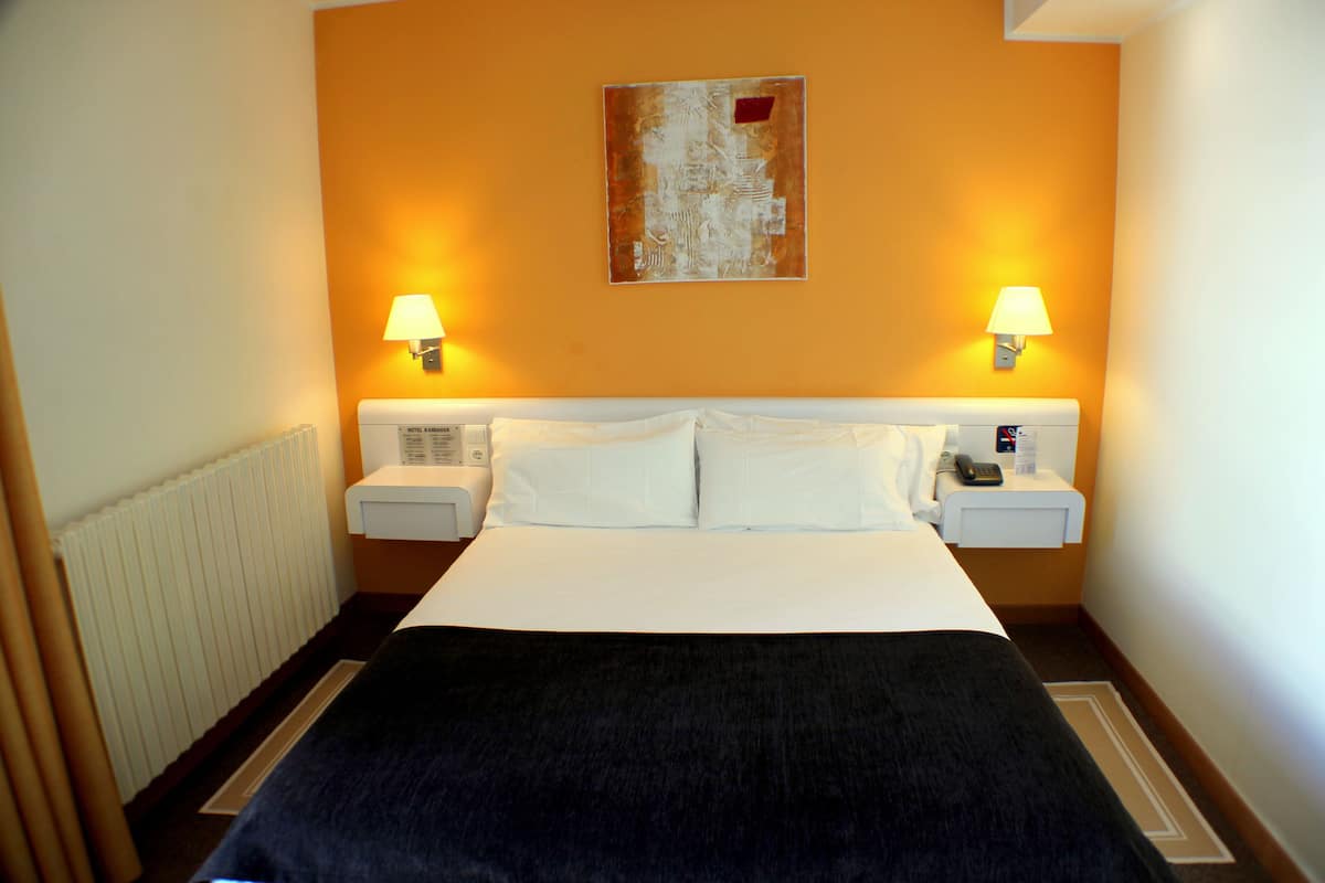 A hotel room with a large bed, two bedside tables with lamps, a radiator, and a painting on the wall.