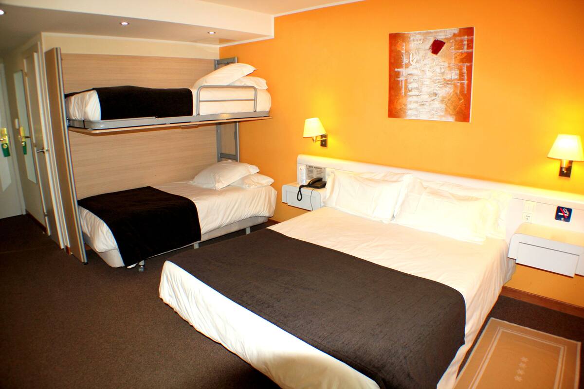 A hotel room with a bunk bed, a single bed, two bedside tables with lamps, a telephone, and a painting on the wall.