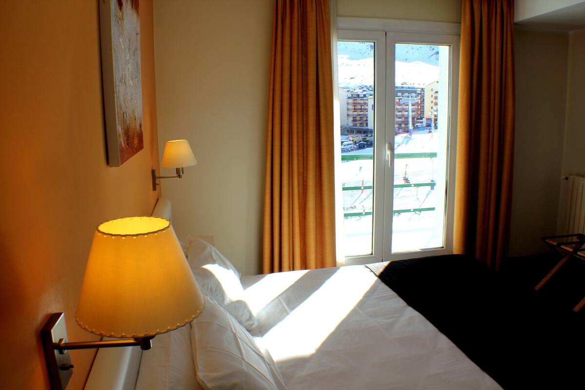 A hotel room with a bed, a lamp, a window with curtains, and a view of buildings outside.