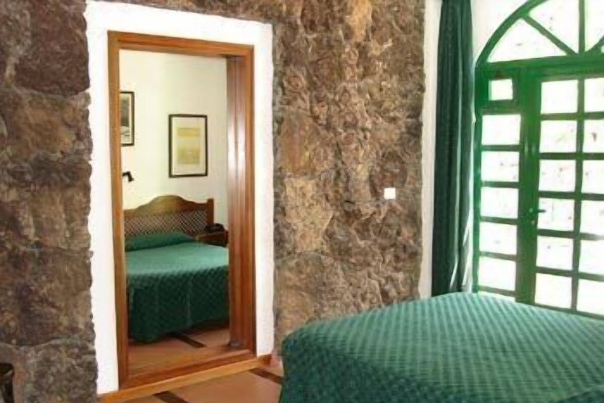 A room with a stone wall, a bed with a green bedspread, a wooden door, and a window with a green frame.