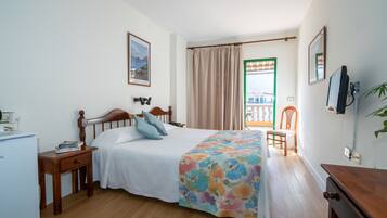 Un dormitorio con cama, escritorio, silla, televisor, ventana con cortinas y vistas al balcón.