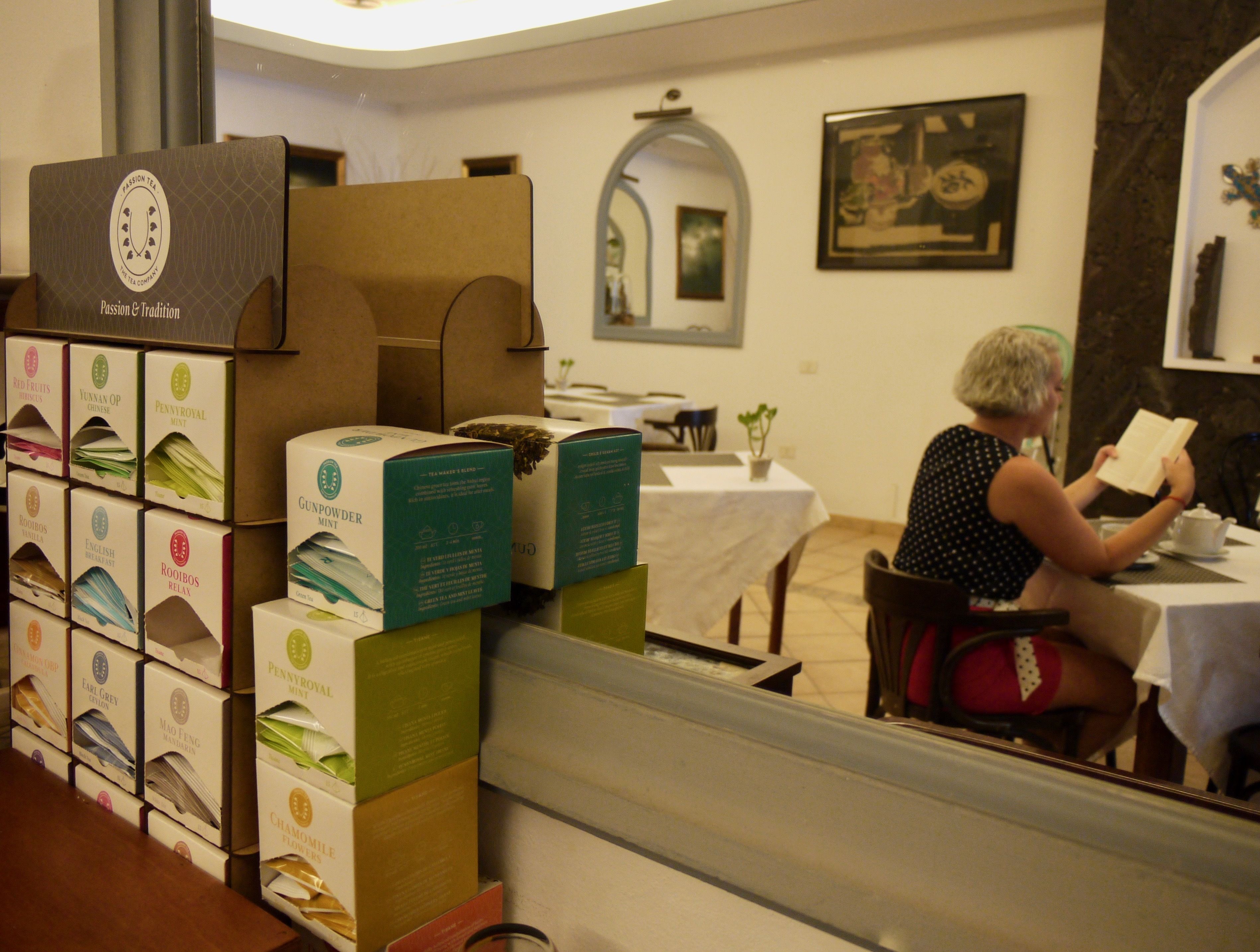 Se exhiben cajas de té en una habitación con una mesa, sillas y un cuadro en la pared. Una persona lee un libro.